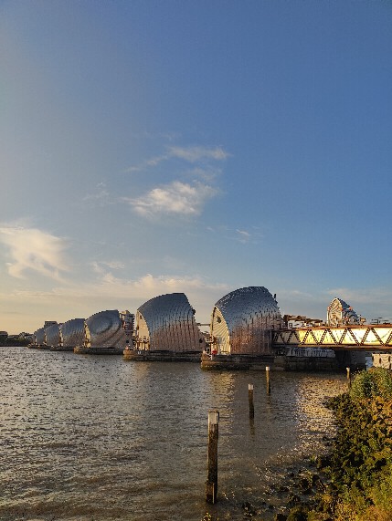 The Thames Barrier in afternoon light from the south bank path, the silver hoods in a line against a blue sky